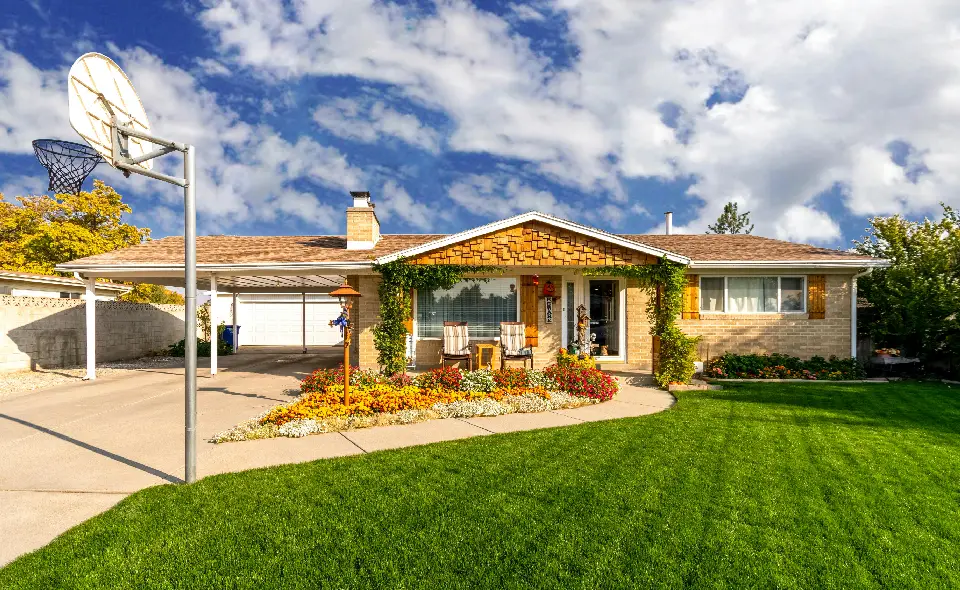 A single-story house with a well-maintained lawn, flower beds, and a basketball hoop in the driveway. The house features a two-car garage and a covered front porch with chairs.